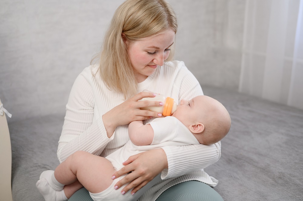Woman tenderly holds small newborn baby in her arms and lovingly feeds him from bottle with an adapted infant formula. Loving mother gives milk to baby and feels happy. Baby care, professional nanny