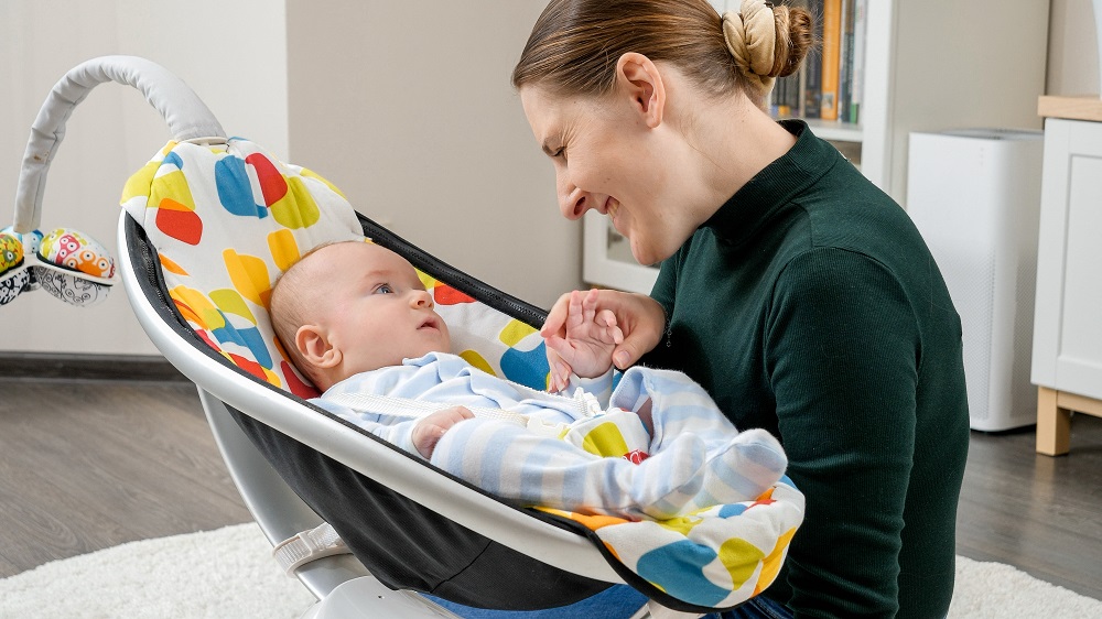 Portrait of smiling mother sitting next to baby son rocking in electric rocker. Child development and childhood